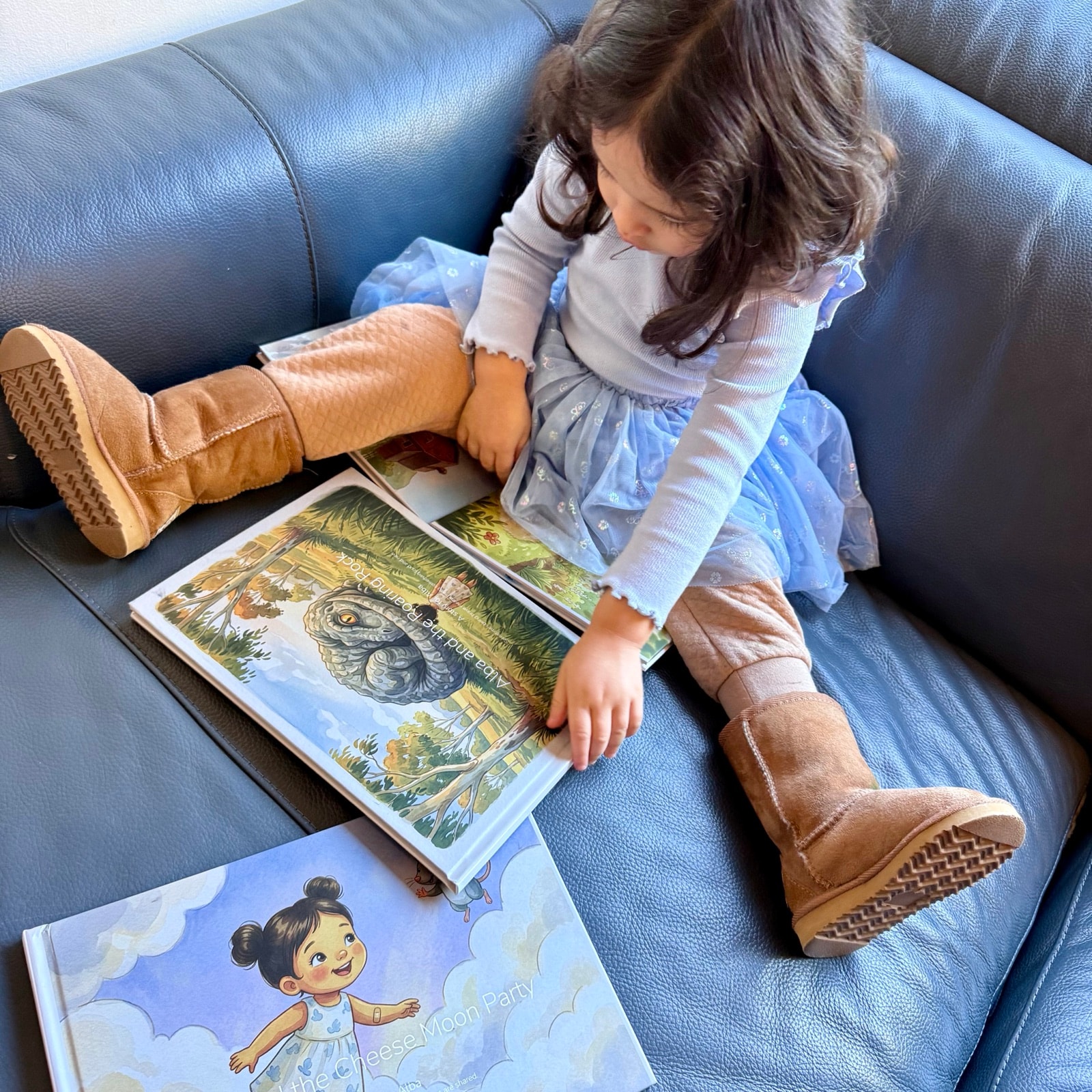 A toddler in a blue dress sits on a couch reading an open Almia personalised hardcover book, with a second personalised book resting beside her.