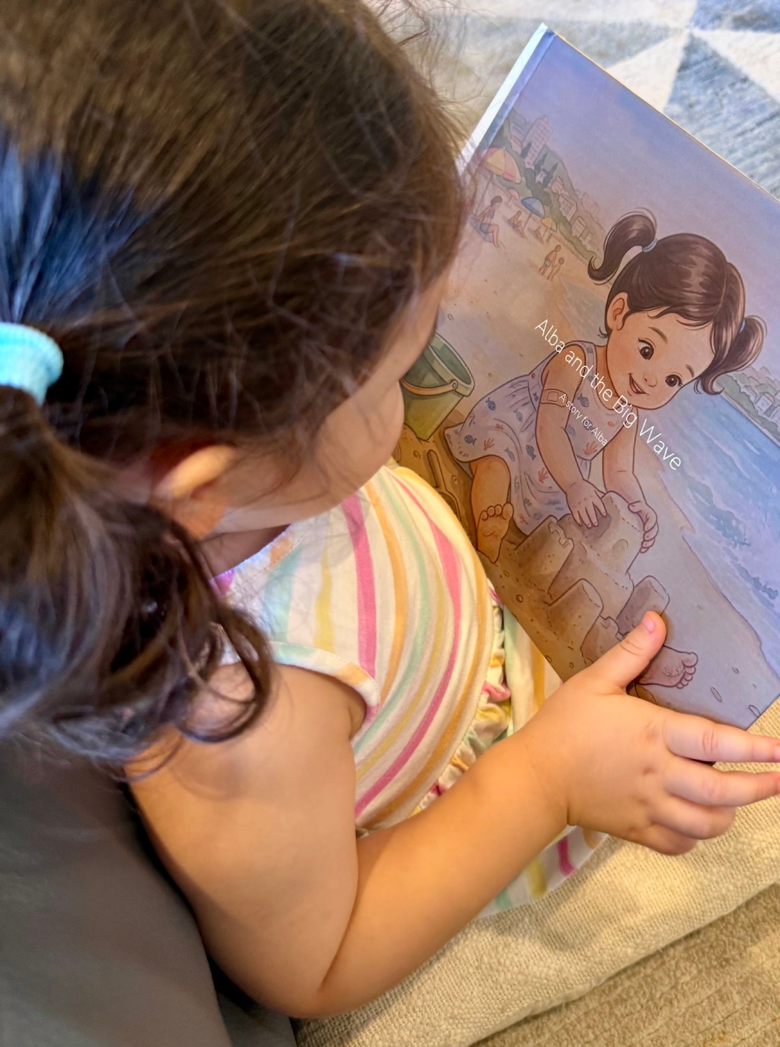 A toddler holding her personalised hardcover, the cover showing her illustrated as the hero of a beach story with a sandcastle.